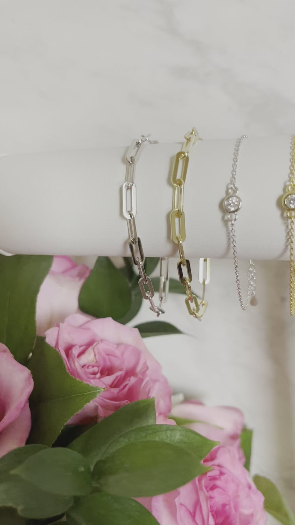 Paperclip chain, single bezel, and evil eye bracelets hanging on white stand.