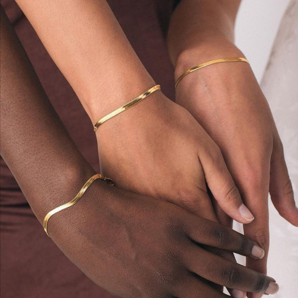 Women of different skin colors holding hands while wearing Herringbone Gold Bracelets. 