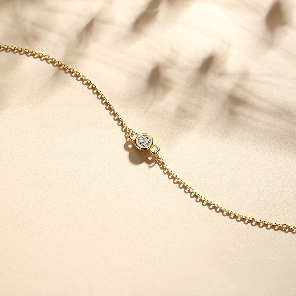18K gold-plated Single Diamond Bezel Bracelet laying flat with flowers as a shadow background.
